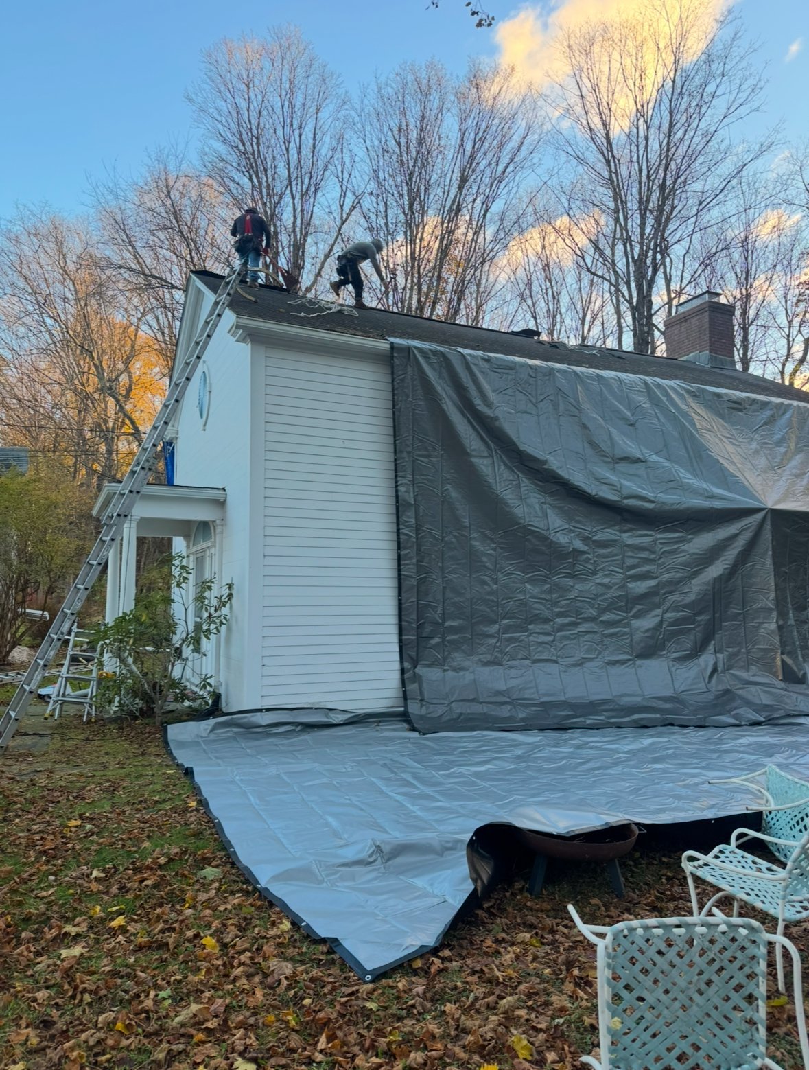 Two workers on a ladder covering a house roof with large dark protective sheeting during renovation, bare trees in background