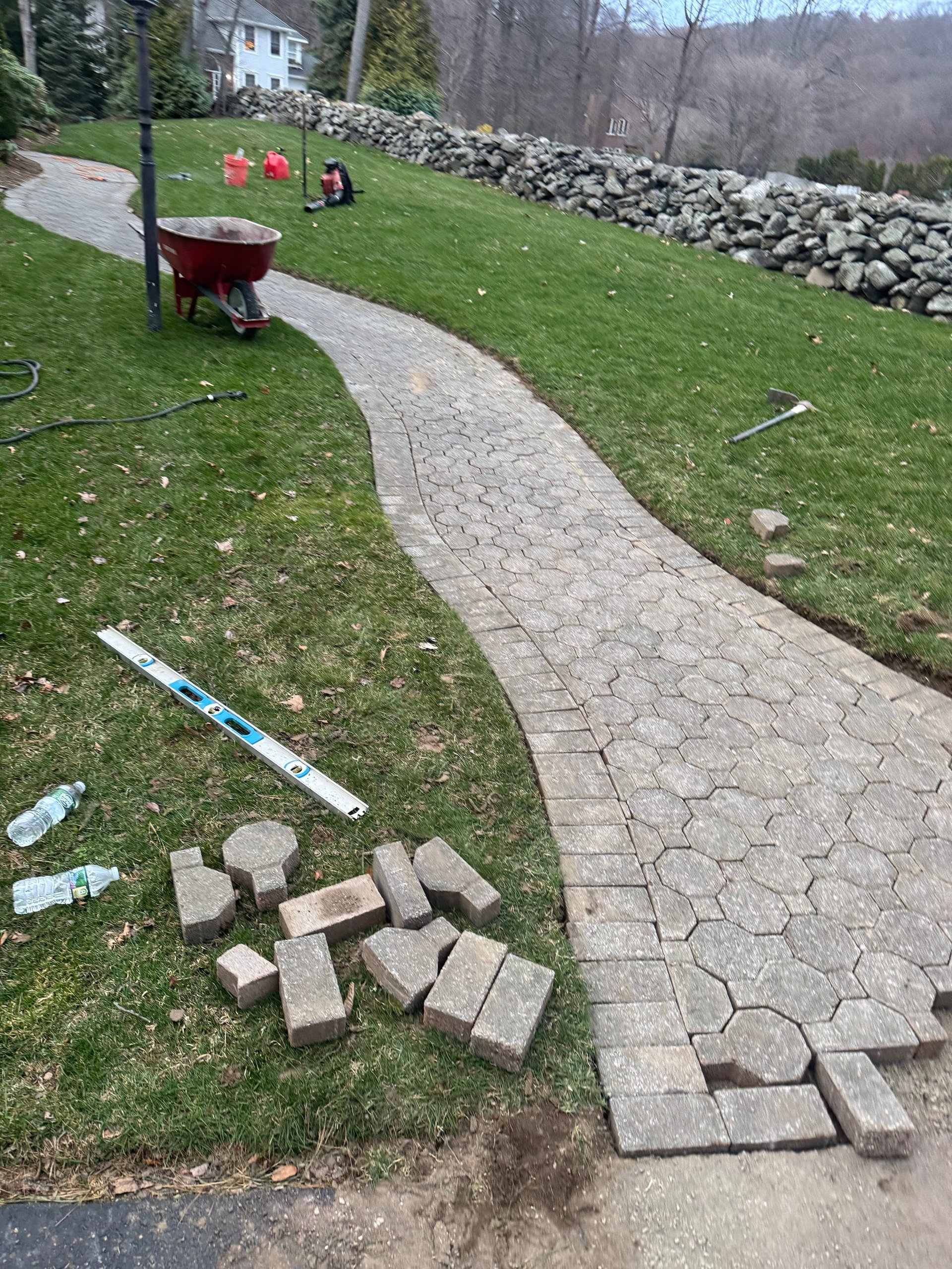 Backyard landscape project with curved paver pathway under construction, loose bricks on grass, wheelbarrow, and stone wall in background