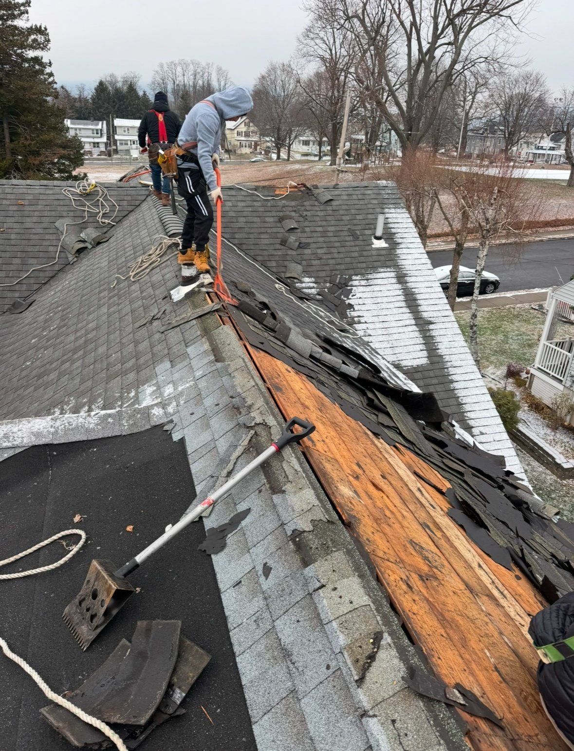 Workers replacing roof shingles on a residential house, showing exposed wood decking and safety equipment
