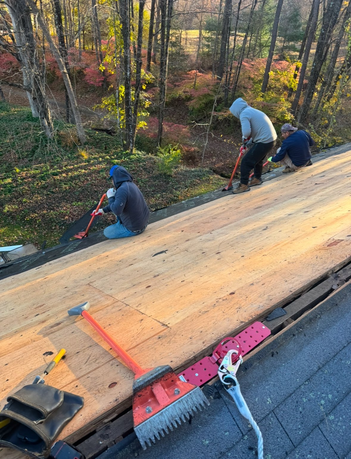 Three workers installing wooden roof shingles on a house with forest and autumn foliage visible in the background