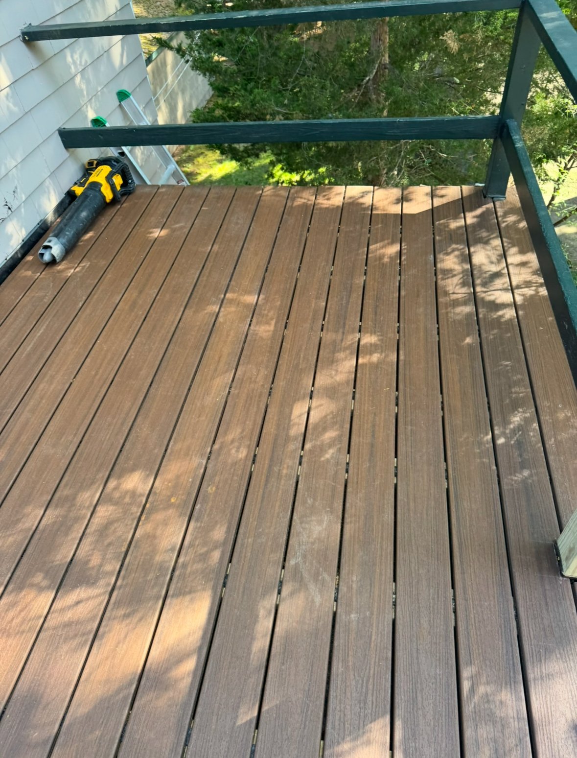 Wooden deck with dark green railing and a yellow power drill on the boards with trees visible in the background