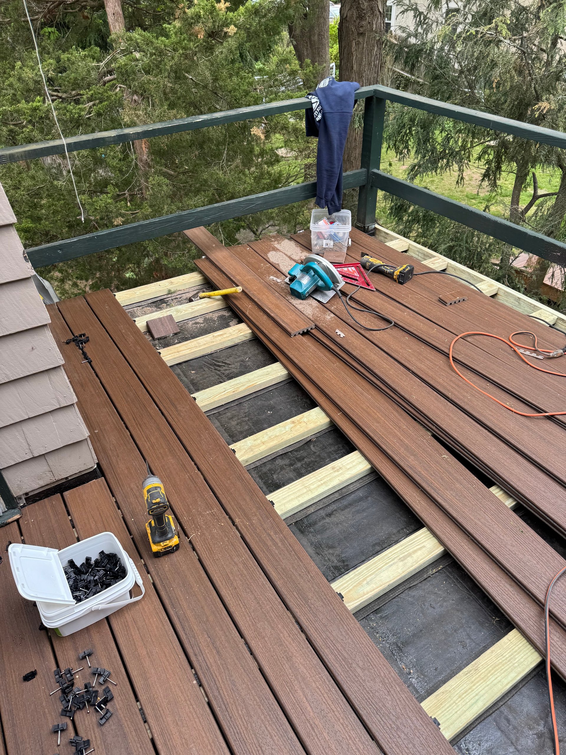 Deck construction in progress with wooden boards, exposed joists, and tools scattered on the surface overlooking a forest