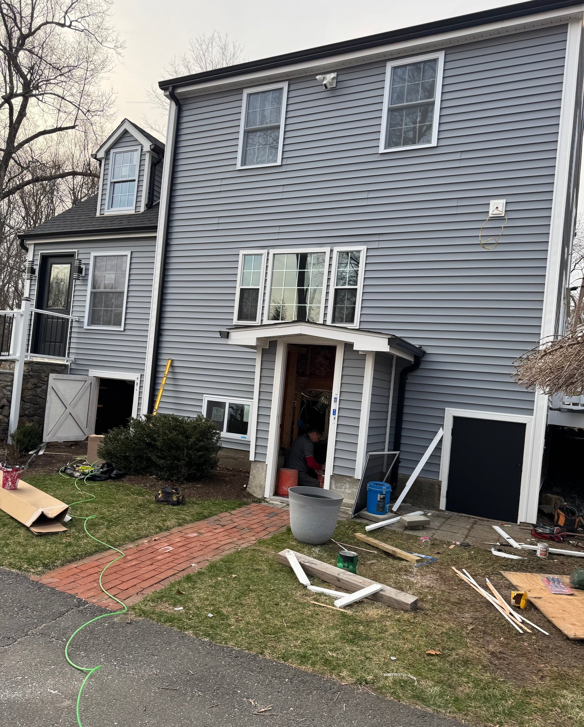 Two-story gray house with deck and backyard under renovation, scattered construction materials and tools visible on lawn