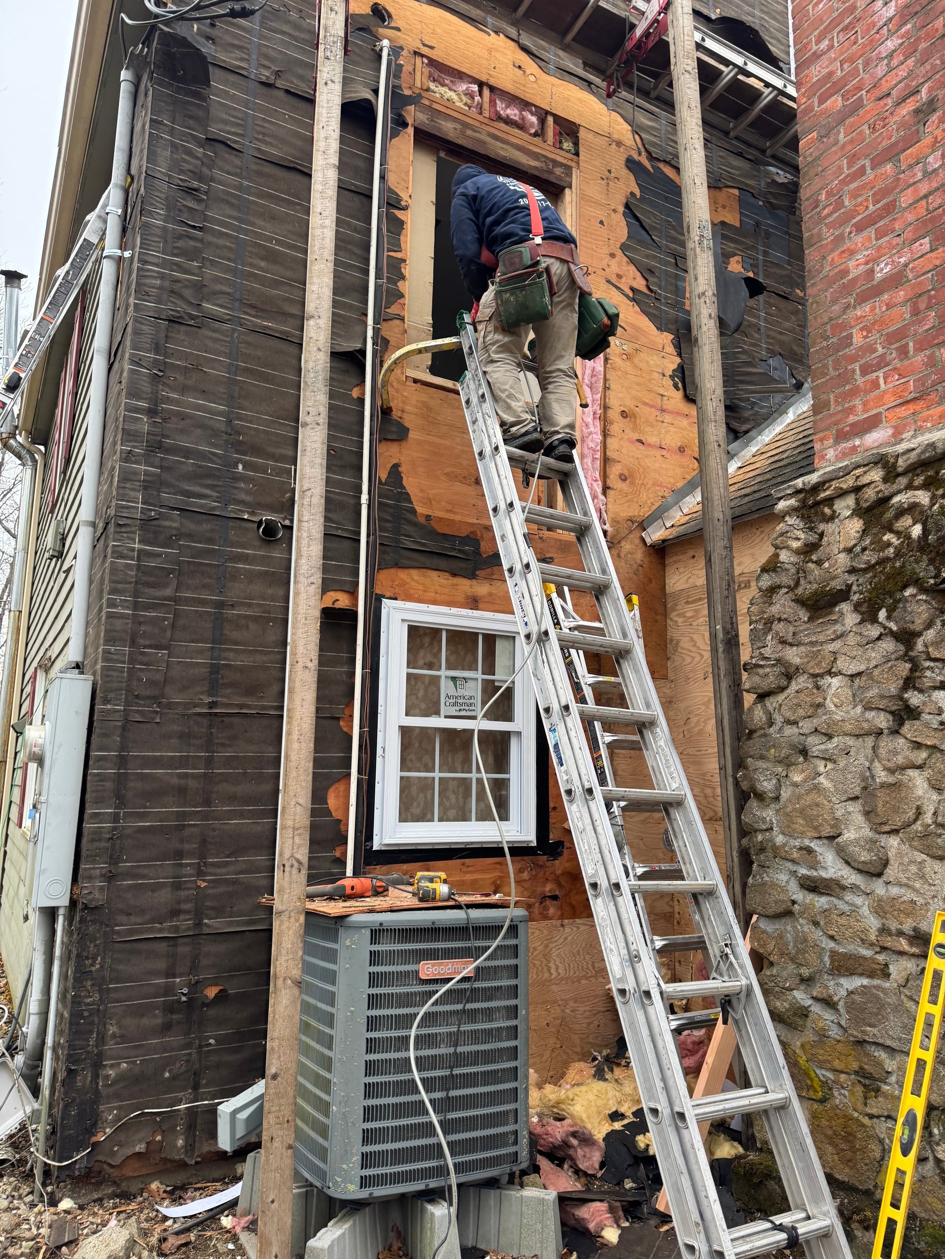 Worker on ladder installing window in wooden wall frame of brick building exterior