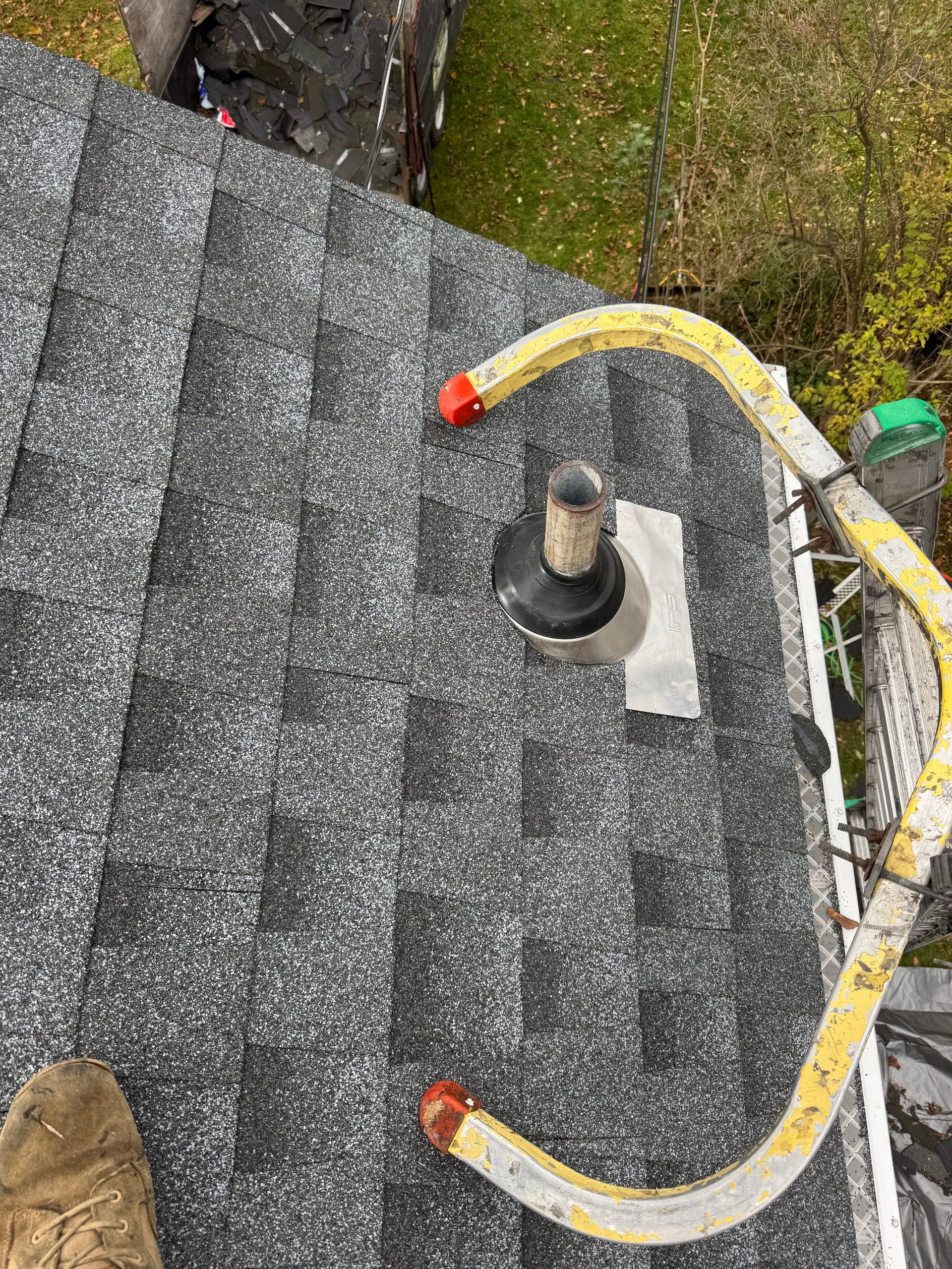 Asphalt shingle roof with yellow safety rope, ladder, and metal vent pipe during maintenance work
