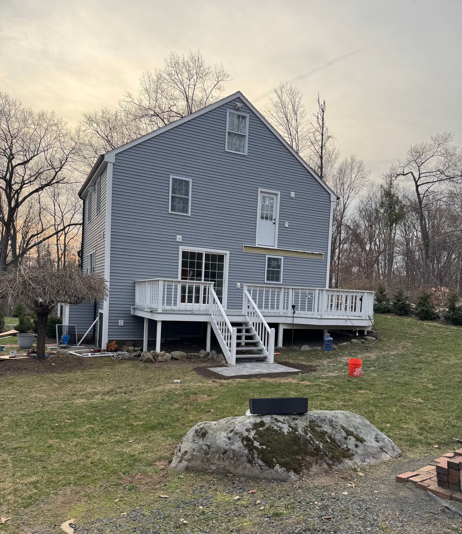 Blue-gray elevated house with white deck and stairs surrounded by bare trees and grass in rural area