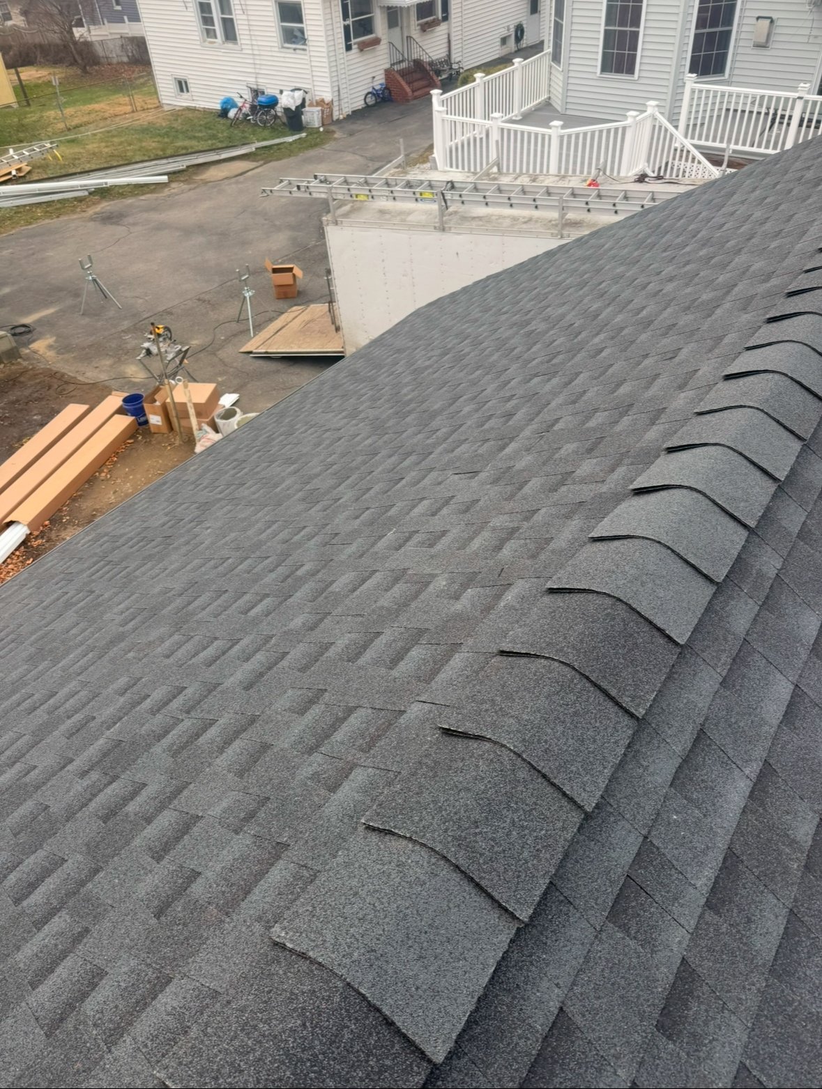 Roofing work in progress with gray shingles laid out on roof, construction materials and workers visible in residential yard below