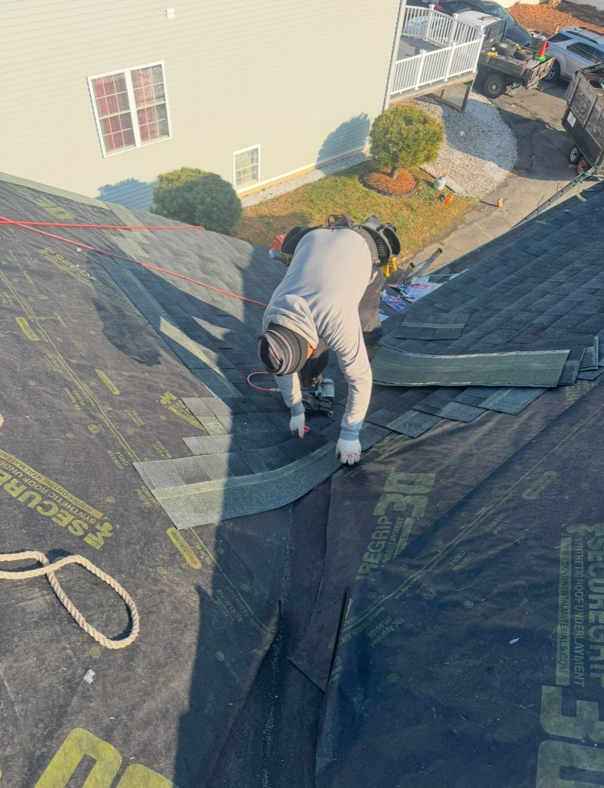 Roofer in white shirt working on asphalt roof shingles with white rope for safety, aerial view of residential property