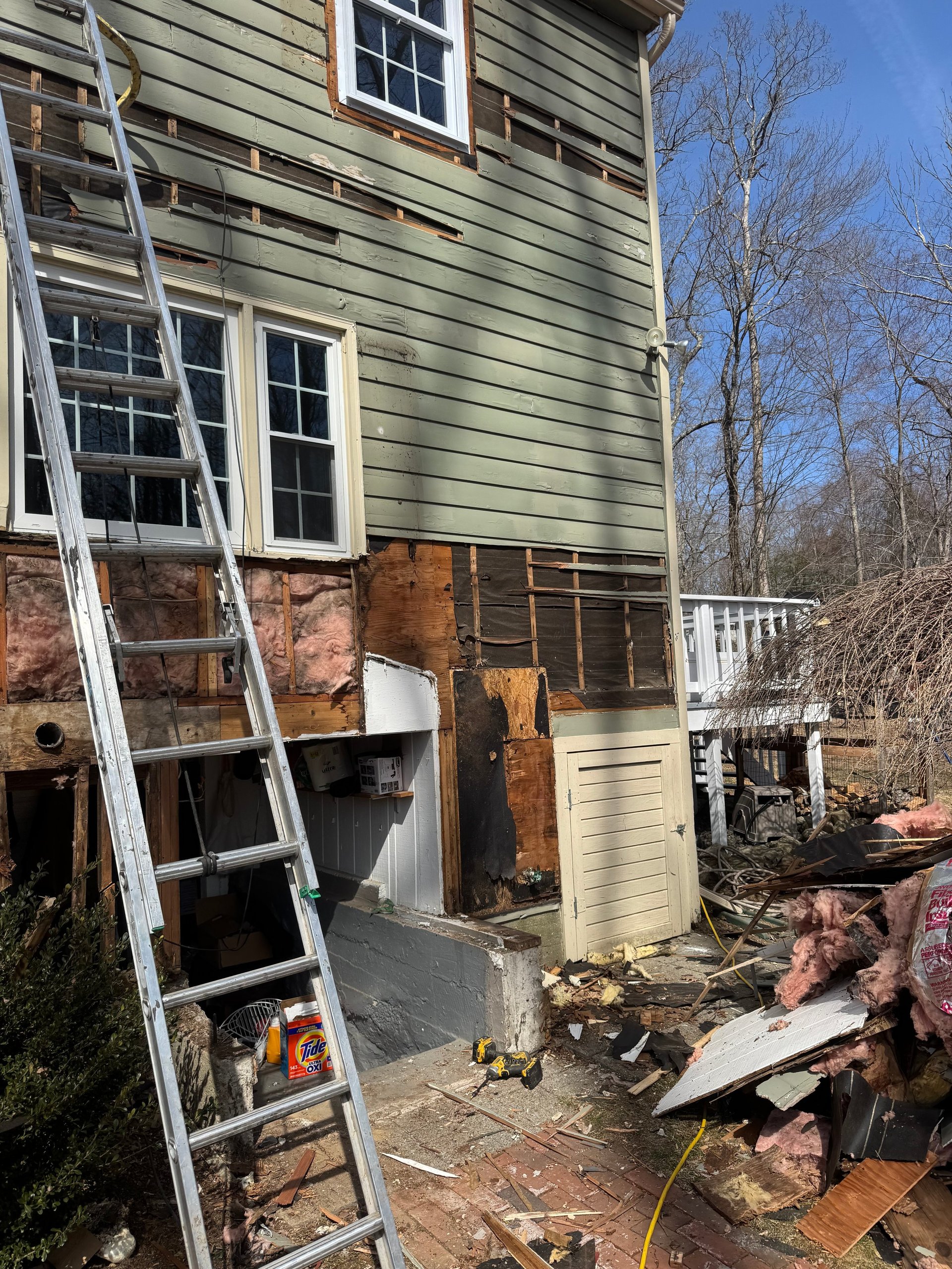 House exterior undergoing renovation with ladder against siding, exposed brick, and construction materials scattered on ground